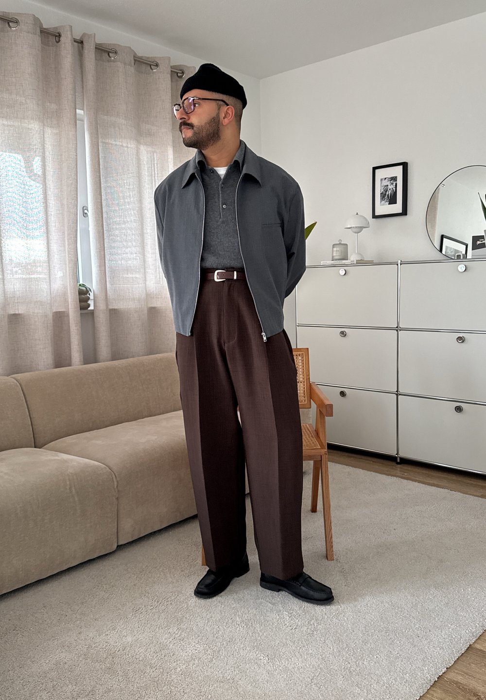 Vyras, dėvintis juodą kepurę, akinius, pilką striukę, tamsiai pilką marškinį, rudus kelnes ir juodus batus, stovi moderniame svetainėje.