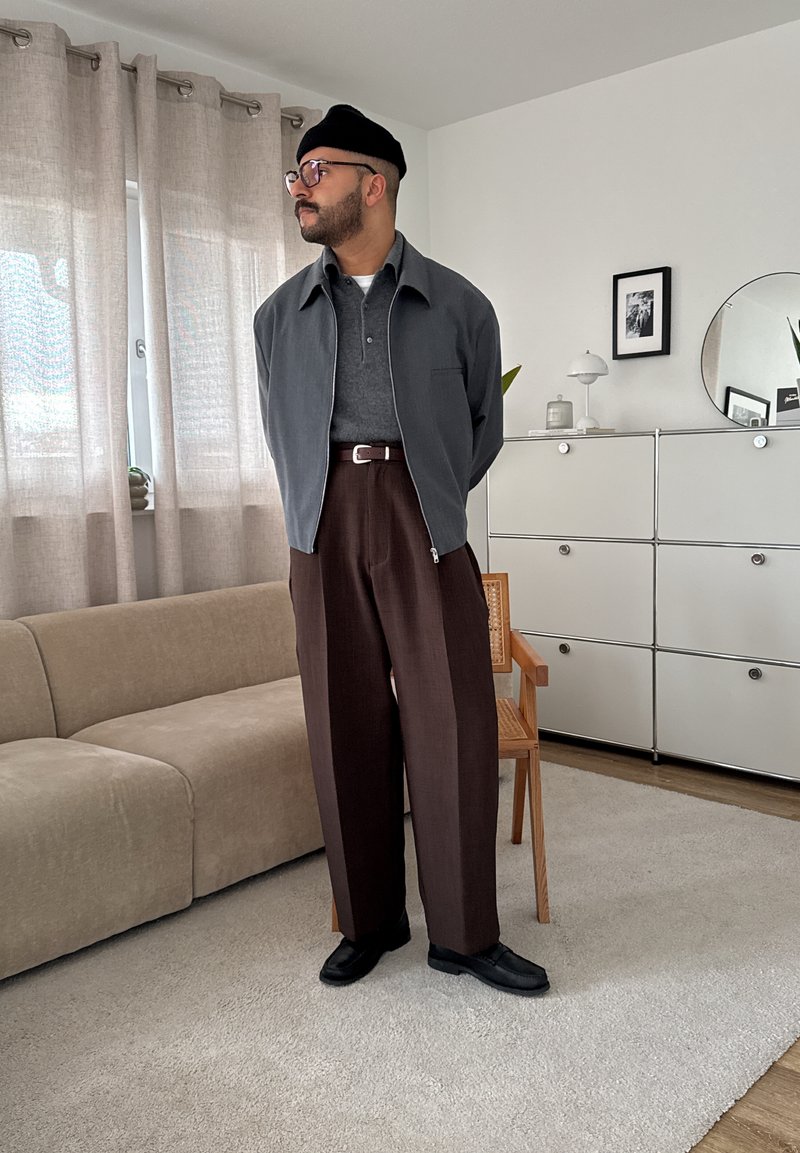 Mand iført en sort beanie, briller, grå jakke, mørkegrå shirt, brune bukser og sorte sko, står i et moderne stue.