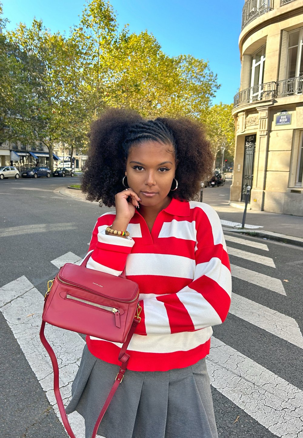 Röd och vit randig polo, grå kjol, håller en liten röd läderväska med dragkedjedetaljer, står på ett övergångsställe med träd i bakgrunden.