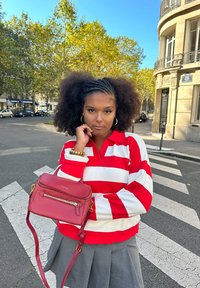 Röd och vit randig polo, grå kjol, håller en liten röd läderväska med dragkedjedetaljer, står på ett övergångsställe med träd i bakgrunden.