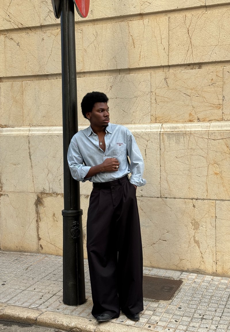 Svetlo modra srajca s spelanim ovratnikom in vezenim detajlom, združena s širokimi črnimi hlačami. Tekstura je gladka, kroj pa sproščen.