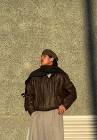 Giacca di pelle marrone, indumento grigio chiaro, sciarpa scura e un basco marrone. Sfondo strutturato. Il soggetto guarda verso l'alto appoggiato al muro.