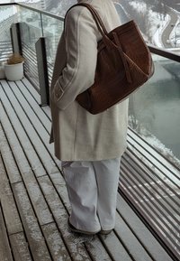 Personne portant un manteau beige et un pantalon blanc tenant un grand sac marron texturé sur un balcon enneigé avec vue sur une rivière et une route.