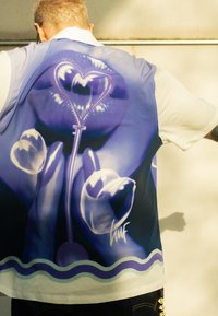 Košile s krátkým rukávem s modro-fialovým grafikou rtů a rukou, s motivem srdce. Vyrobená z lehké tkaniny s hladkou texturou.