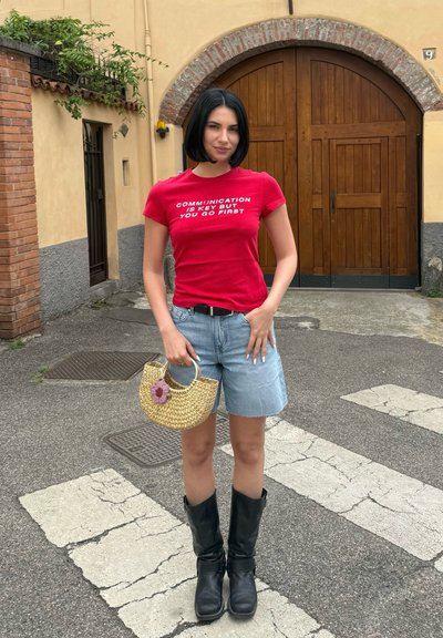 Mujer con el pelo corto y oscuro lleva una camiseta roja con texto blanco, pantalones cortos vaqueros, botas negras y sostiene un pequeño bolso de paja en el exterior.