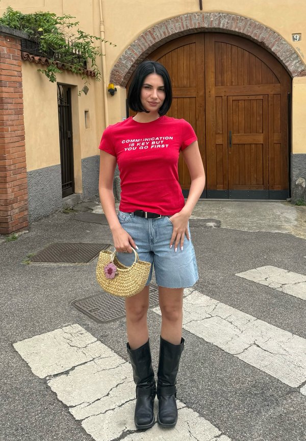 Vrouw met kort donker haar draagt een rood shirt met witte tekst, een jeansshort, zwarte laarzen en houdt buiten een klein strooien tasje vast.