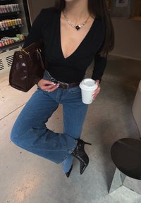 Top nero a maniche lunghe con scollo a v, jeans blu a vita alta, borsa in pelle marrone e tacchi neri appuntiti, che tiene in mano una tazza di caffè bianca.