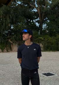 Idrottare i en åtsittande svart New Balance t-shirt och svarta mönstrade Adidas-byxor, med solglasögon och en basebollkeps, står utomhus.