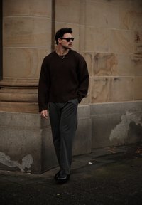 Donkerbruine oversized trui, grijze broek en zwarte schoenen. Zonnebril en gouden ketting benadrukken de outfit. Gladde textuur en ontspannen pasvorm.