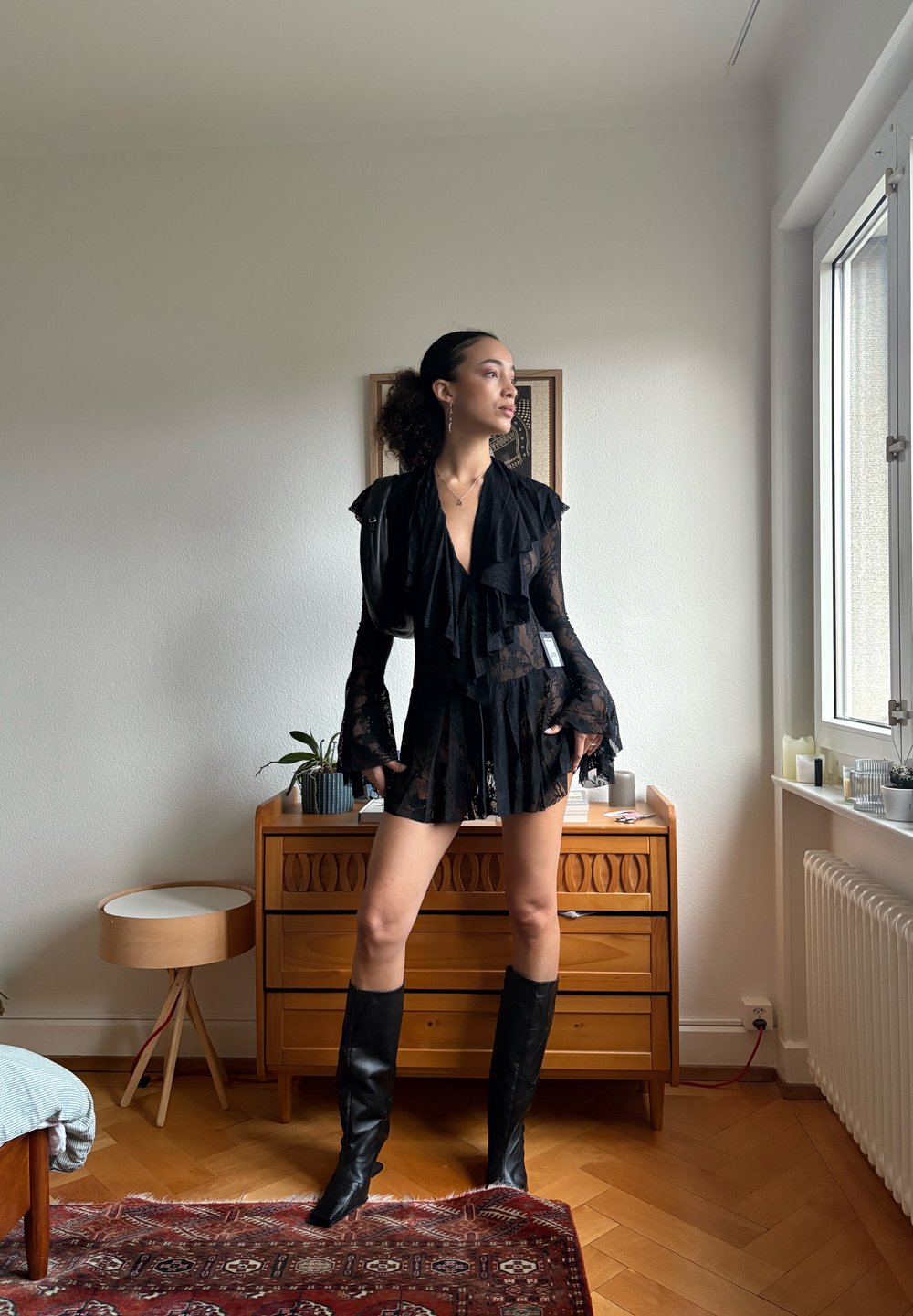 Combinaison noire en dentelle avec manches volantées et décolleté en V profond, associée à des bottes noires montantes jusqu'au genou. Cadre intérieur avec mobilier et décoration en bois.