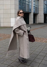 Manteau en laine beige avec un design drapé et des détails à franges, assorti d'un sac à main bordeaux et de chaussures à talons kitten foncés. Tenant une tasse de café.