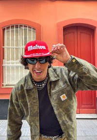 Rød baseballcaps med "Mahou" brodert, kamuflasjemønstret jakke, svart skjorte, lagdelte sølvkjeder, mørke solbriller og et lekent uttrykk.