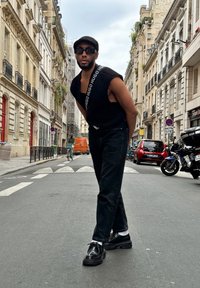 Μαύρο αμάνικο πουκάμισο με λεπτομέρειες λογότυπου, μαύρα τζιν, γυαλιστερά μαύρα παπούτσια και λευκές κάλτσες, ποζάρει σε έναν αστικό δρόμο με κτίρια στο φόντο.