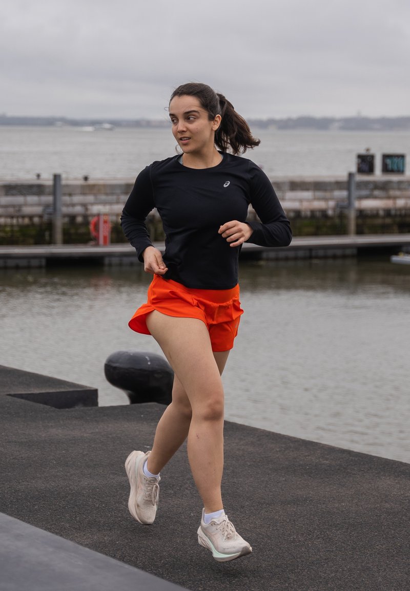 Μαύρη αθλητική μπλούζα με μακρύ μανίκι και πορτοκαλί σορτσάκι τρεξίματος; φοράει ανοιχτόχρωμα αθλητικά παπούτσια; τρέχει κατά μήκος ενός παραλιακού μονοπατιού με γκρι ουρανό στο φόντο.