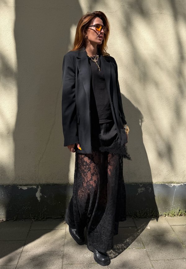 Žena v čiernom saku, čiernej čipkovej sukni, čiernych čižmách, jantárových slnečných okuliaroch a vrstvených zlatých náhrdelníkoch stojí pri slnkom osvietenú stenu.