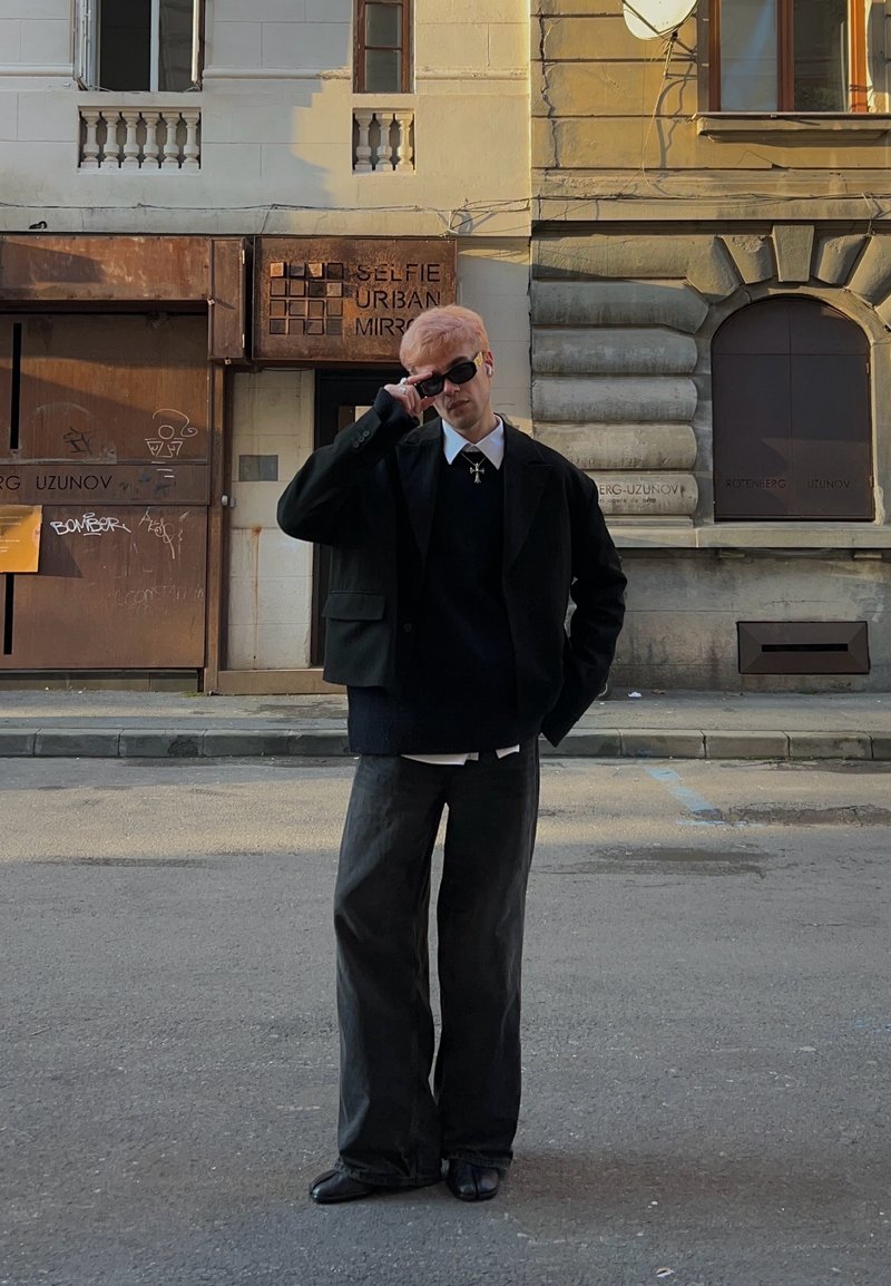 Veste noire oversize, pull bleu marine, chemise blanche à col, jean ample foncé et chaussures noires. Cheveux courts rose clair, portant des lunettes de soleil.