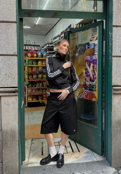 Chaqueta negra con cremallera y rayas blancas, pantalones cortos negros hasta la rodilla, calcetas blancas y zapatos negros. De pie en la puerta de una tienda.