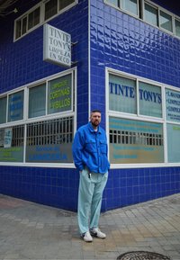 Casaco de fleece azul, calças azul-claro e sapatilhas brancas. O fundo apresenta uma parede de azulejos azuis com sinalização em espanhol sobre serviços de lavandaria.