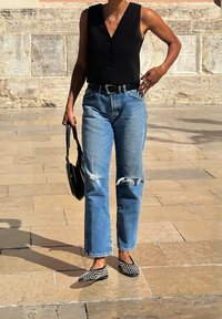 Top senza maniche nero con bottoni, jeans strappati azzurri chiari, cintura nera, borsa nera e scarpe slip-on nere con pois bianchi.