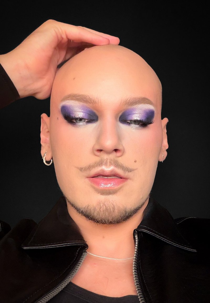 Personne chauve avec un maquillage des yeux violet et argenté, des lèvres brillantes, une fine moustache et une barbiche, portant des boucles d'oreilles et une veste noire, main touchant la tête.