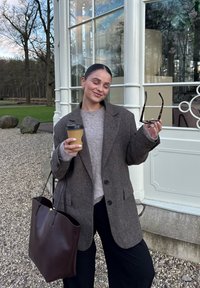 Bruine wollen jas over een grijze trui, met een koffiecup en een zonnebril. Bruine leren tote bag. Grindgrond en groen in de achtergrond.