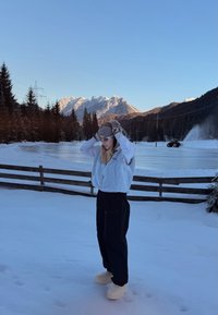 Personne portant des vêtements d'hiver ajuste son chapeau en se tenant sur un sol enneigé près d'une clôture en bois, avec des montagnes et des arbres en arrière-plan.