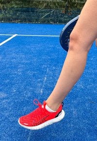 Červená sportovní bota s texturovaným svrškem a bílou podrážkou, nošená s bílou ponožkou. Pozadí zobrazuje modrý povrch kurtu.