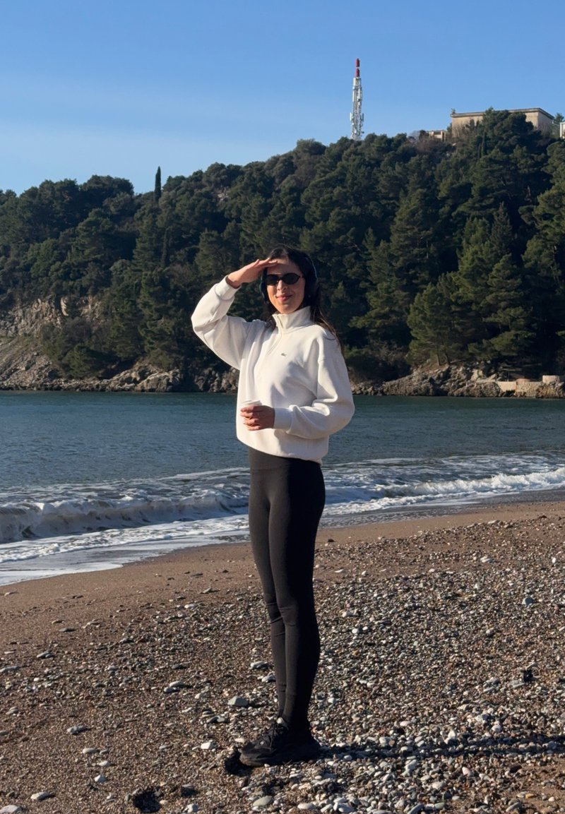 Femme en veste blanche et leggings noirs, se protégeant les yeux du soleil et tenant une tasse, debout sur une plage rocheuse avec des vagues et une colline boisée en arrière-plan.