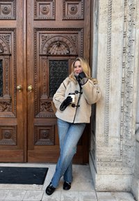 Bézs, polárbéléses kabát fekete szegéllyel, szürke pulóver, kék, bővülő szárú farmernadrág, fekete mokaszinek; fehér csészét tart, egy fából készült ajtón támaszkodik.