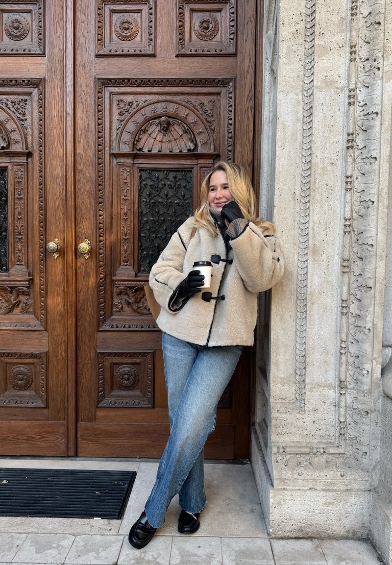 Beiger, mit Fleece gefütterter Mantel und schwarzer Besatz, grauer Pullover, blaue Schlagjeans, schwarze Loafers; hält eine weiße Tasse und lehnt an einer Holztür.
