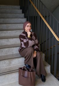 Pulover tricotat cu model de diamante maro și bej, pantaloni maro cu picioare largi, cizme închise la culoare, accesorii aurii și o geantă tote maro pe scările din beton.