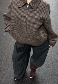 Braune Hahnentritt-Reißverschlussjacke mit großem Kragen, kombiniert mit weit geschnittenen schwarzen Hosen und burgunderfarbenen Stiefeln mit Absatz. Strukturierter Stoff und lockere Passform.