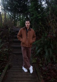 Veste isolante marron avec un col sombre, chemise noire en dessous, pantalon marron et baskets blanches, sur fond de forêt verte.