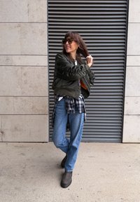 Veste bomber vert olive, t-shirt blanc, chemise à carreaux nouée autour de la taille, jean bleu et bottes marron. Debout contre un mur à lamelles.