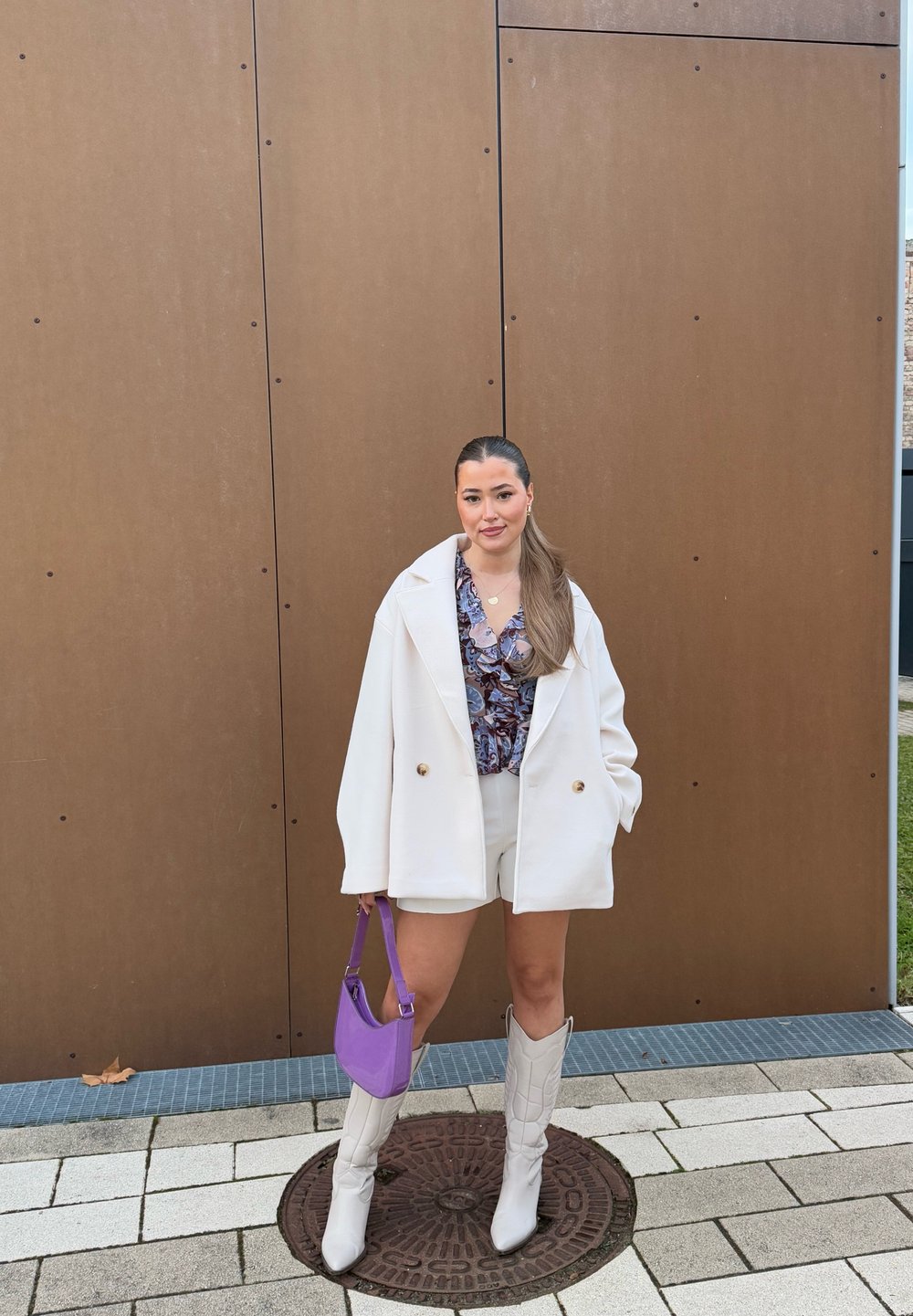 Übergroßer weißer Blazer, gemusterte Bluse, weiße Shorts, weiße kniehohe Stiefel und eine kleine lila Tasche vor einem braunen strukturierten Hintergrund.