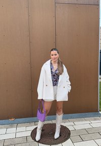 Übergroßer weißer Blazer, gemusterte Bluse, weiße Shorts, weiße kniehohe Stiefel und eine kleine lila Tasche vor einem braunen strukturierten Hintergrund.