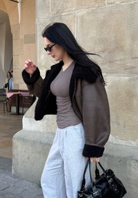 Casaco de pele castanha com colarinho e punhos de pele preta, blusa cinza justa, calças largas cinza claro, mala preta, óculos de sol pretos e elegantes.
