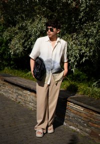 Προσωπικά κοντομάνικο ελαφρύ πουκάμισο με σχέδιο βουνών, συνδυασμένο με μπεζ παντελόνι φαρδύ και γκρι παντόφλες. Κρατάει μαύρη τσάντα.