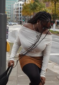 Top de manga comprida, com decote ombro a ombro e riscas pretas e brancas, combinado com uma saia de camurça castanha e collants pretos, segurando uma mala.