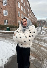 Jeune femme portant une capuche grise et un manteau en fourrure blanche à pois, croisant les bras et se tenant dans une rue urbaine enneigée avec des bâtiments en briques en arrière-plan.