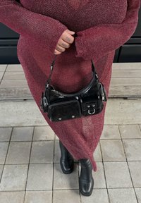 Čierna kabelka s bezpečnostným zipsom, lesklou textúrou a striebornými prvkami. Nosená s dlhými, bordovými, textúrovanými šatami a čiernymi čižmami.