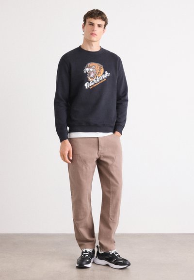 Sudadera color navy con gráfico de un león y texto, combinada con pantalones beige y zapatillas negras. Ajuste casual, material de algodón, puños de canalé.