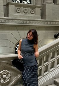 Femme avec lunettes portant un haut gris sans manches et une jupe à pois, appuyée sur un escalier en pierre orné à l'intérieur d'un bâtiment historique.