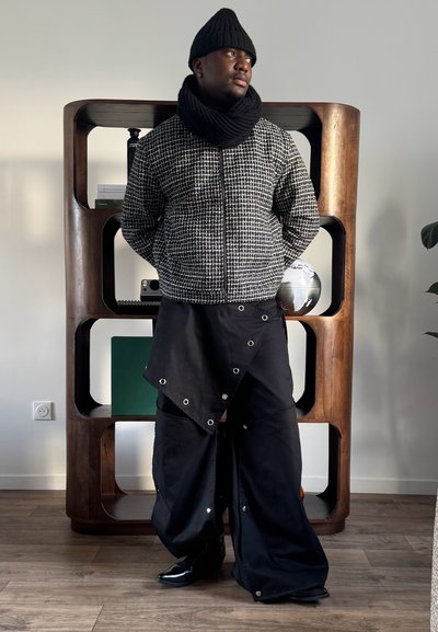 Hombre con gorro de punto negro y bufanda, chaqueta a cuadros, pantalones anchos negros con detalles de presión, de pie en el interior frente a una estantería de madera con libros y un globo terráqueo.