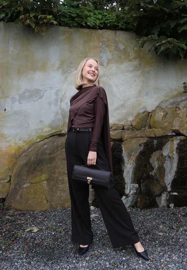 Mujer con el pelo corto y rubio, lleva una camiseta marrón, pantalones negros de pierna ancha, sostiene un bolso negro, de pie sobre grava junto a un muro de piedra.