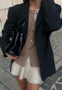 Sort blæser over en beige strikket cardigan med knapper, parret med en hvid fuzzy nederdel. Holder en lille sort lædertaske.