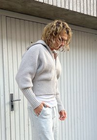 Lys grå strikket cropped genser med glidelås og ribbestruktur samt hette; kombinert med lys grå joggebukser, stående foran en trebakgrunn.