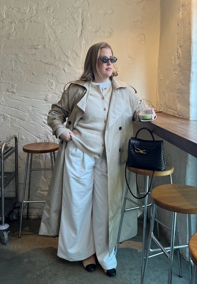 Abrigo trench beige sobre un suéter crema y pantalones anchos blancos. Bolso negro con textura trenzada. Gafas de sol oscuras, vaso en mano.