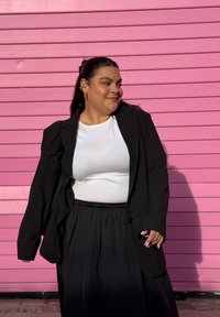 Blazer preto sobre um top branco, combinado com uma saia preta e fluida. O fundo apresenta riscas horizontais cor-de-rosa. Cabelo longo e escuro e brincos visíveis.