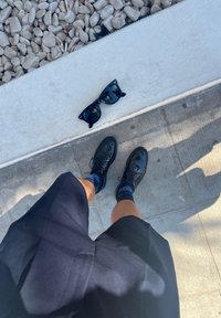 Scarpe in pelle nera e calzini blu scuro sono indossati con pantaloni corti blu navy. Occhiali da sole neri giacciono su una superficie di cemento chiaro accanto a ciottoli grigi.